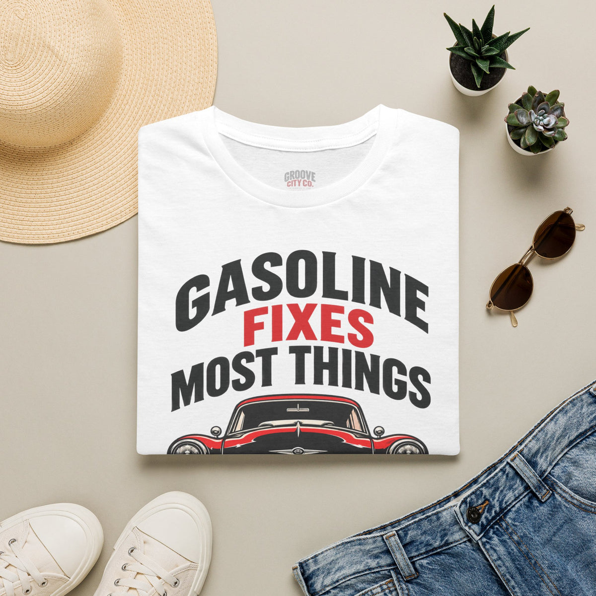 Folded white “Gasoline Fixes Most Things” T-shirt with a red and black classic car design, styled with jeans and sunglasses.