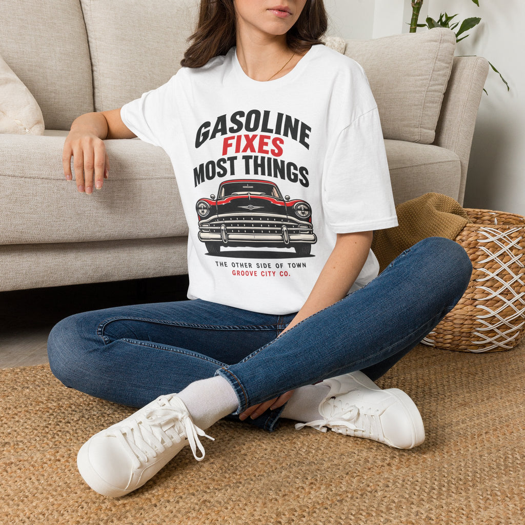 Woman seated indoors wearing a white “Gasoline Fixes Most Things” T-shirt with a red and black classic car graphic.